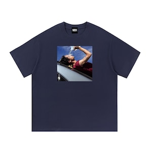 Puede incluir: Una camiseta azul marino de manga corta con un estampado cuadrado que muestra a una persona con camisa granate vertiéndose agua sobre la cabeza bajo un cielo azul. La prenda tiene cuello redondo y una etiqueta negra WRYX en el interior.