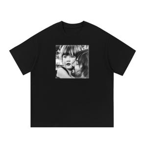 May include: A black short-sleeved t-shirt featuring a square, black and white photographic print on the chest. The graphic depicts a close-up of a young child with bangs looking forward while being embraced by an adult with a beard.