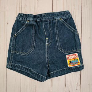 May include: Dark blue denim shorts with an elastic waistband and yellow stitching. Features two front pockets and a patch with the word "THOMAS" and a cartoon train. A children's clothing item.