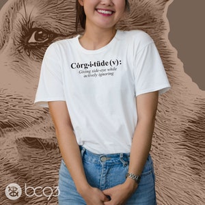 May include: White t-shirt with a humorous definition of "Corg-i-tüde" printed on the front. The definition reads: "Giving side-eye while actively ignoring."