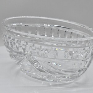 May include: Clear, oval-shaped crystal bowl with a decorative, textured design. The bowl features a series of vertical lines and oval shapes around the rim and a swirling pattern at the base. The bowl is made of clear glass.