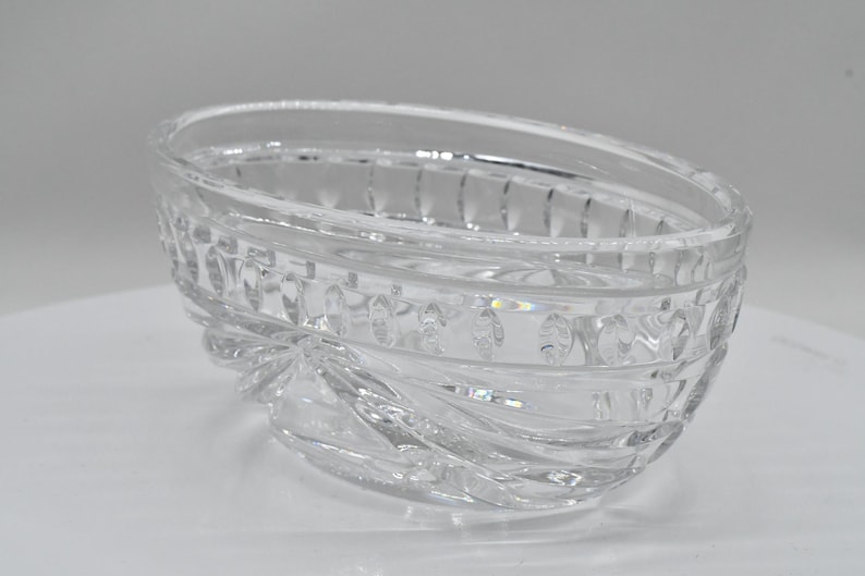 May include: Clear, oval-shaped crystal bowl with a decorative, textured design. The bowl features a series of vertical lines and oval shapes around the rim and a swirling pattern at the base. The bowl is made of clear glass.