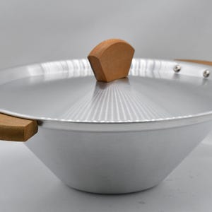 May include: A silver wok with a matching lid and wooden handles. The lid has a wooden handle on top. The wok is made of metal and has a wide, sloped design. The handles are made of light-colored wood.