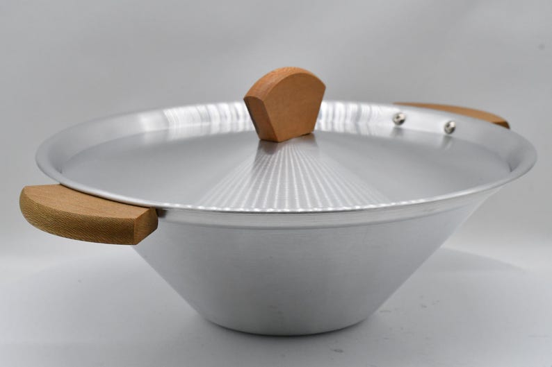 May include: A silver wok with a matching lid and wooden handles. The lid has a wooden handle on top. The wok is made of metal and has a wide, sloped design. The handles are made of light-colored wood.
