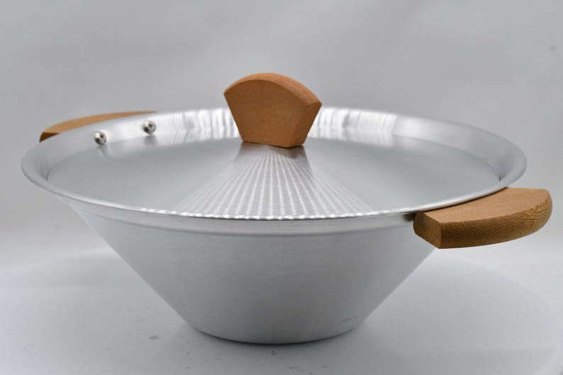 May include: A silver-colored wok with a matching lid and wooden handles. The lid has a wooden handle on top. The wok is designed with a wide, sloped bowl and is suitable for cooking various Asian-inspired dishes.