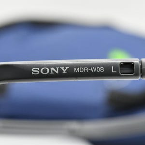 May include: Close-up of a gray Sony MDR-W08 headphone arm. The text "SONY MDR-W08 L" is printed in white on the arm. The background is blurred, with a blue and green color scheme.