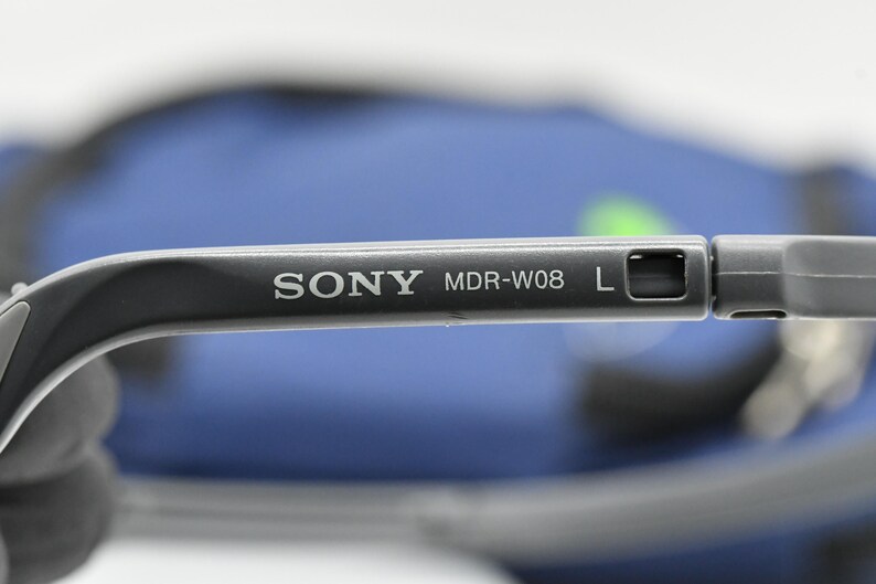 May include: Close-up of a gray Sony MDR-W08 headphone arm. The text "SONY MDR-W08 L" is printed in white on the arm. The background is blurred, with a blue and green color scheme.