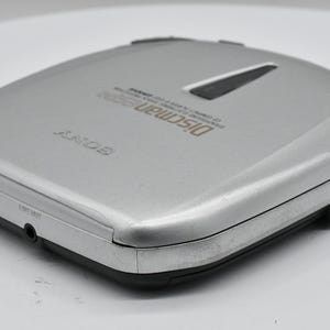 May include: Silver Sony Discman D-200 CD player. The portable CD player has a rectangular shape with rounded corners. The top has a black accent and the words "Discman D-200" printed in gold. It has a headphone jack and other ports.