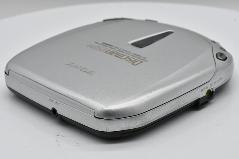May include: Silver Sony Discman D-200 CD player. The portable CD player has a rectangular shape with rounded corners. The top has a black accent and the words "Discman D-200" printed in gold. It has a headphone jack and other ports.