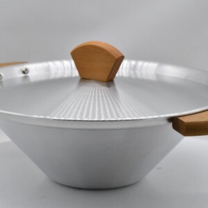 May include: A silver-colored wok with a matching lid and wooden handles. The lid features a wooden handle. The wok is designed with a wide, sloped bowl and is suitable for cooking various dishes.