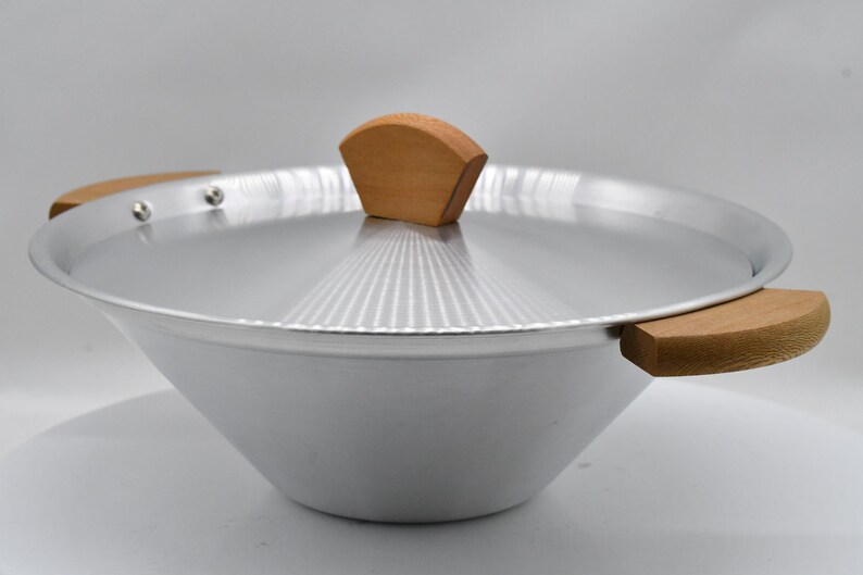 May include: A silver-colored wok with a matching lid and wooden handles. The lid features a wooden handle. The wok is designed with a wide, sloped bowl and is suitable for cooking various dishes.