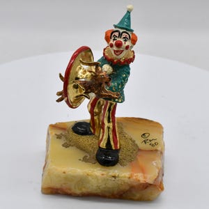 May include: A whimsical clown figurine with a green jacket, red and gold striped pants, and a red and gold horn. The clown is holding a small brown animal and a gold and red object. The figurine is mounted on a rectangular, tan-colored base.