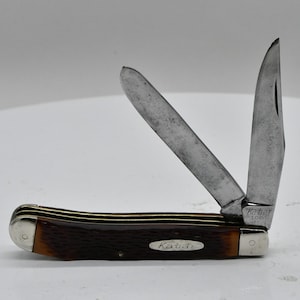 May include: A brown and silver pocket knife with two open blades. The handle is a dark brown color with a silver end cap. The word "Kabar" is printed on the handle and the blade.