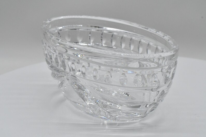 May include: Clear, oval-shaped crystal bowl with a decorative, textured design. The bowl features a series of oval and rectangular patterns around the rim and sides. The crystal bowl is a decorative item.