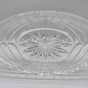 May include: Clear, oval-shaped crystal dish with intricate etched patterns. The center features a starburst design, surrounded by curved lines and floral accents. The dish is made of transparent glass, reflecting light.