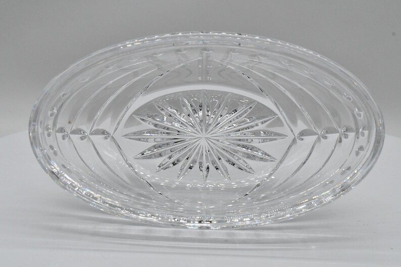 May include: Clear, oval-shaped crystal dish with intricate etched patterns. The center features a starburst design, surrounded by curved lines and floral accents. The dish is made of transparent glass, reflecting light.