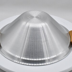 May include: A silver-colored metal serving dish with a conical shape and vertical ridges. It has two wooden handles and sits on a white, circular base. The dish is designed for food service.