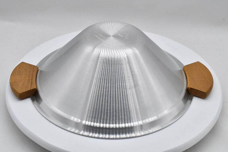 May include: A silver-colored metal serving dish with a conical shape and vertical ridges. It has two wooden handles and sits on a white, circular base. The dish is designed for food service.