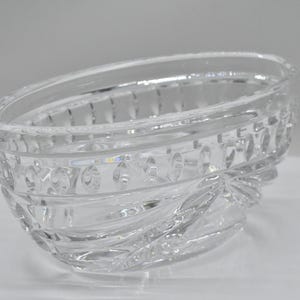 May include: A clear, oval-shaped glass bowl with a decorative, textured design. The bowl features a series of horizontal bands with repeating patterns and a wavy, textured base. The glass is transparent, allowing light to pass through.