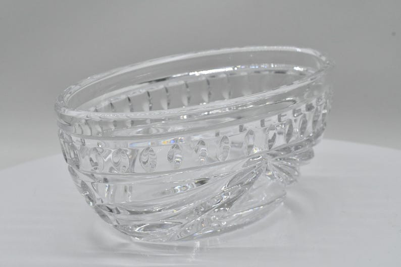 May include: A clear, oval-shaped glass bowl with a decorative, textured design. The bowl features a series of horizontal bands with repeating patterns and a wavy, textured base. The glass is transparent, allowing light to pass through.
