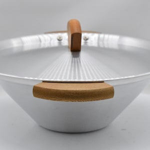 May include: A silver wok with a matching lid and wooden handles. The wok has a wide, conical shape and a lid with a wooden handle. The handles are made of light-colored wood. The wok is designed for cooking.