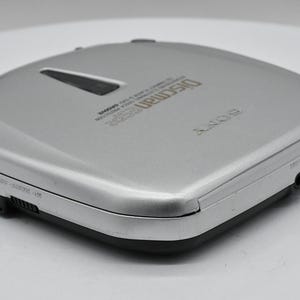 May include: Silver Sony Discman portable CD player. The device has a rectangular shape with rounded edges and a black base. The top of the player has the word "Discman" printed on it.