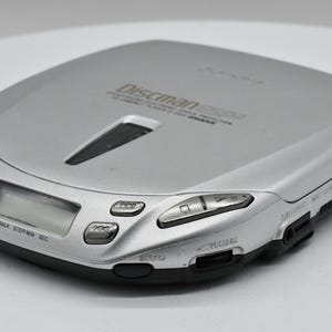 May include: Silver Discman ESP2 portable CD player. The device has a rectangular shape with rounded edges and a black base. It features control buttons, a display screen, and a volume control. The text "Discman ESP2" is visible on the top.