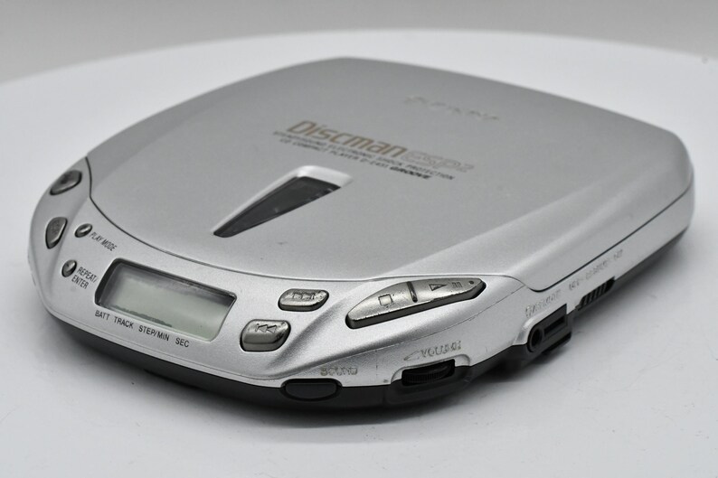May include: Silver Discman ESP2 portable CD player. The device has a rectangular shape with rounded edges and a black base. It features control buttons, a display screen, and a volume control. The text "Discman ESP2" is visible on the top.