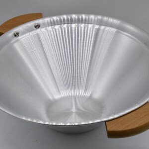 May include: A silver metal bowl with a brushed finish and two wooden handles. The bowl has a wide, shallow design and is likely used for serving or display. The wooden handles are attached with small silver rivets.