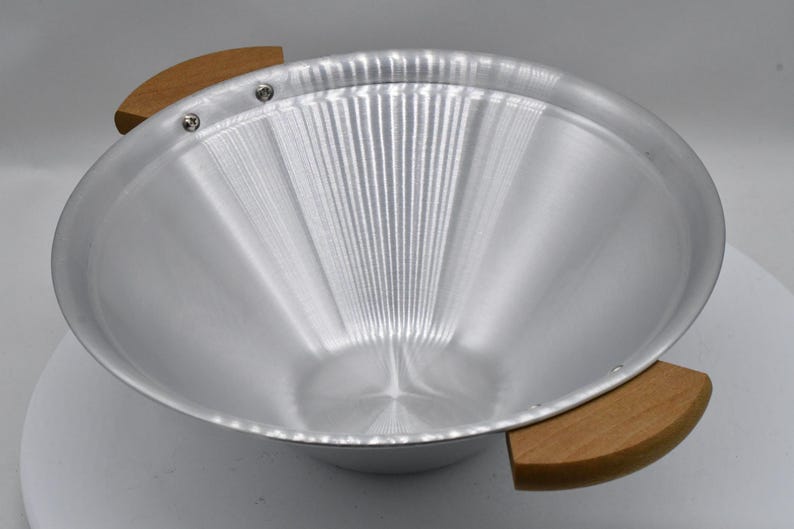 May include: A silver metal bowl with a brushed finish and two wooden handles. The bowl has a wide, shallow design and is likely used for serving or display. The wooden handles are attached with small silver rivets.