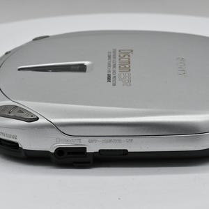 May include: Silver Sony Discman ESP2 CD player. The portable CD player has a rounded rectangular shape with buttons and a headphone jack. The text "Discman ESP2" is printed on the top. The device is silver and black.