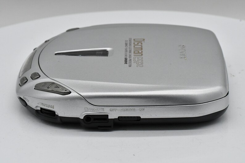 May include: Silver Sony Discman ESP2 CD player. The portable CD player has a rounded rectangular shape with buttons and a headphone jack. The text "Discman ESP2" is printed on the top. The device is silver and black.