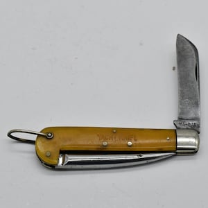 May include: A vintage folding pocket knife with a yellow handle and a partially open silver blade. The text "YACHT KNIFE" is visible on the handle. The blade is marked "KA-BAR".