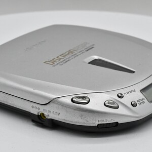 May include: Silver Discman portable CD player with a black base. The device has several buttons, a digital display, and the text "Discman" printed on the top. The player is designed to play compact discs.