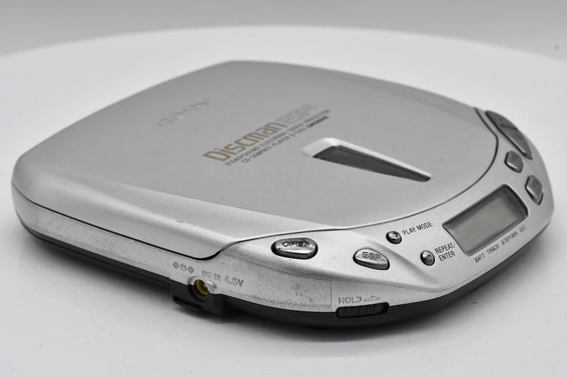 May include: Silver Discman portable CD player with a black base. The device has several buttons, a digital display, and the text "Discman" printed on the top. The player is designed to play compact discs.