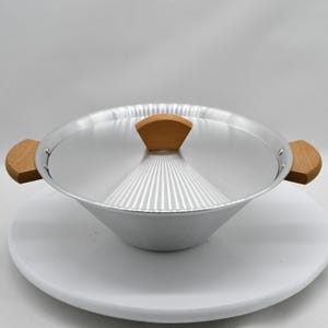 May include: A silver-colored cooking pot with a matching lid and wooden handles. The pot has a conical shape and is sitting on a white surface. The lid has a wooden handle. The pot is designed for cooking.