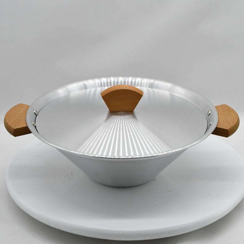 May include: A silver-colored cooking pot with a matching lid and wooden handles. The pot has a conical shape and is sitting on a white surface. The lid has a wooden handle. The pot is designed for cooking.
