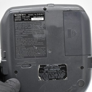 May include: A gray Sony Discman D-E451, with a battery compartment and various labels. The labels include the Sony logo, model number, serial number, and certification information. The back of the device is visible.