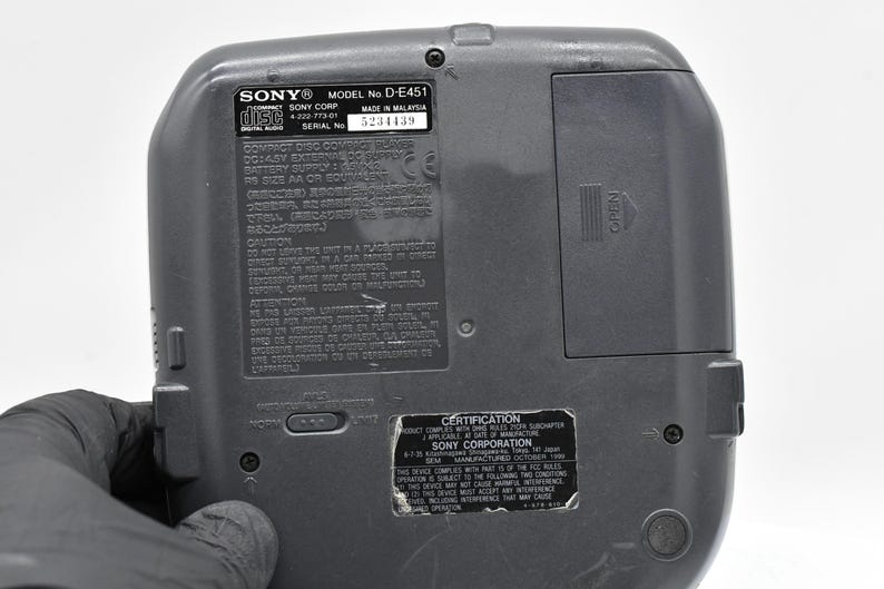 May include: A gray Sony Discman D-E451, with a battery compartment and various labels. The labels include the Sony logo, model number, serial number, and certification information. The back of the device is visible.