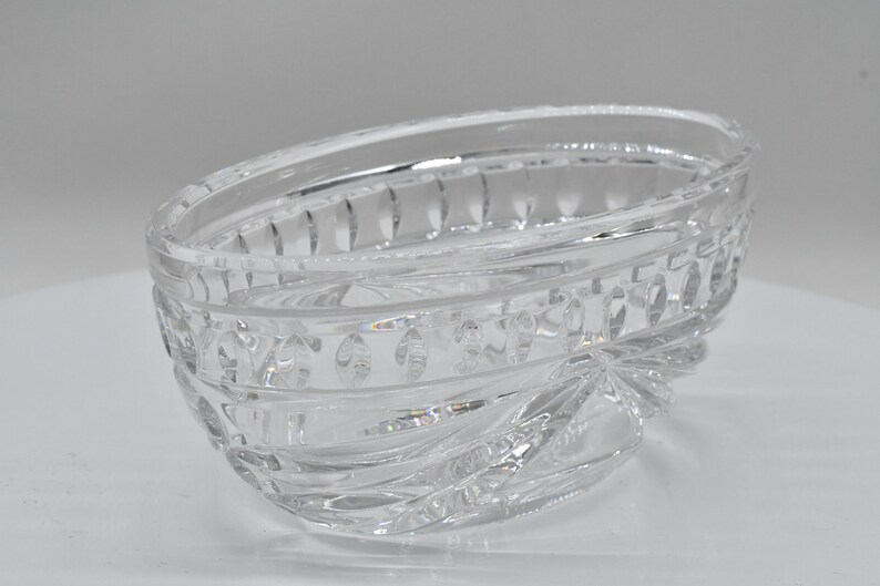 May include: Clear, oval-shaped crystal bowl with intricate cut glass detailing. The bowl features a decorative pattern around the rim and a starburst design at the base. A decorative piece for serving or display.