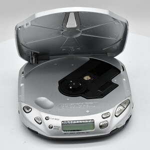 May include: Silver portable CD player with the lid open. The player has several buttons and a small display screen. The CD tray is black. The device is on a white surface.