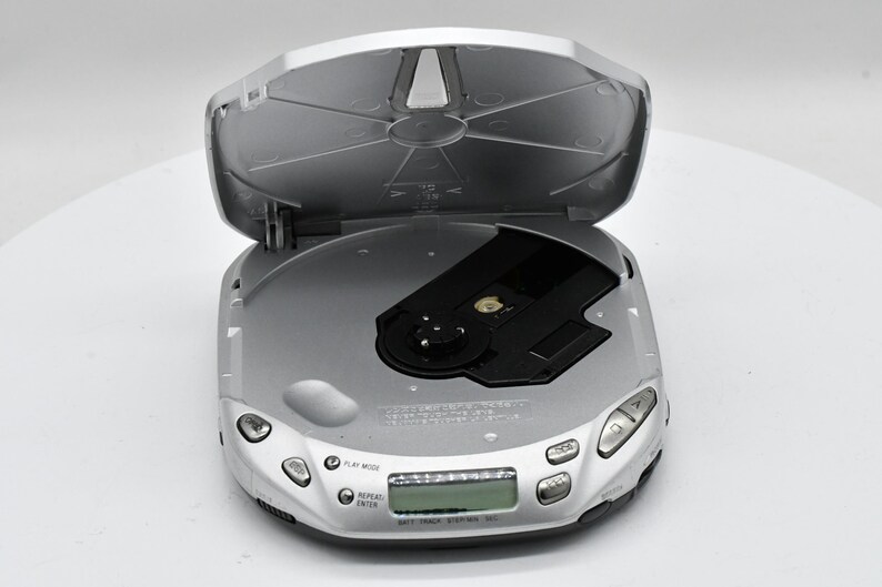 May include: Silver portable CD player with the lid open. The player has several buttons and a small display screen. The CD tray is black. The device is on a white surface.