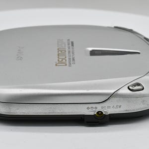 May include: Silver Sony Discman portable CD player. The device has a rectangular shape with rounded edges and a black base. The top surface has the word "SONY" and "Discman" printed on it, along with other text and buttons.