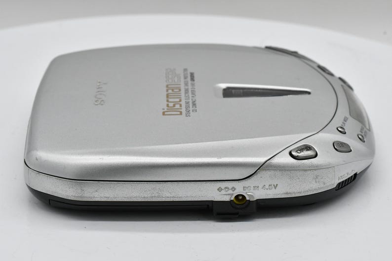 May include: Silver Sony Discman portable CD player. The device has a rectangular shape with rounded edges and a black base. The top surface has the word "SONY" and "Discman" printed on it, along with other text and buttons.