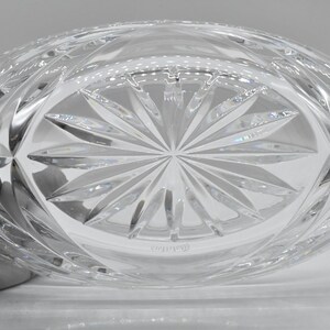 May include: Clear, oval-shaped crystal bowl with a detailed, geometric pattern. The base features a starburst design, and the sides have a repeating diamond pattern. The bowl is held by a gloved hand, showcasing the intricate craftsmanship of the crystal.