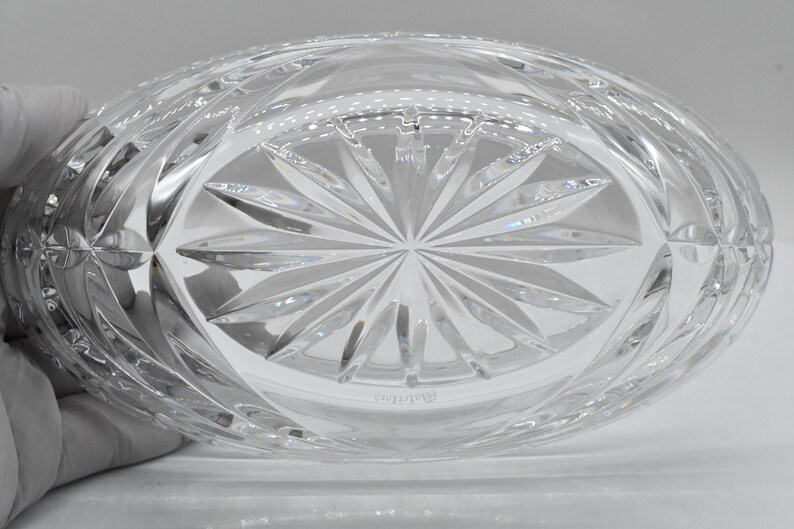 May include: Clear, oval-shaped crystal bowl with a detailed, geometric pattern. The base features a starburst design, and the sides have a repeating diamond pattern. The bowl is held by a gloved hand, showcasing the intricate craftsmanship of the crystal.