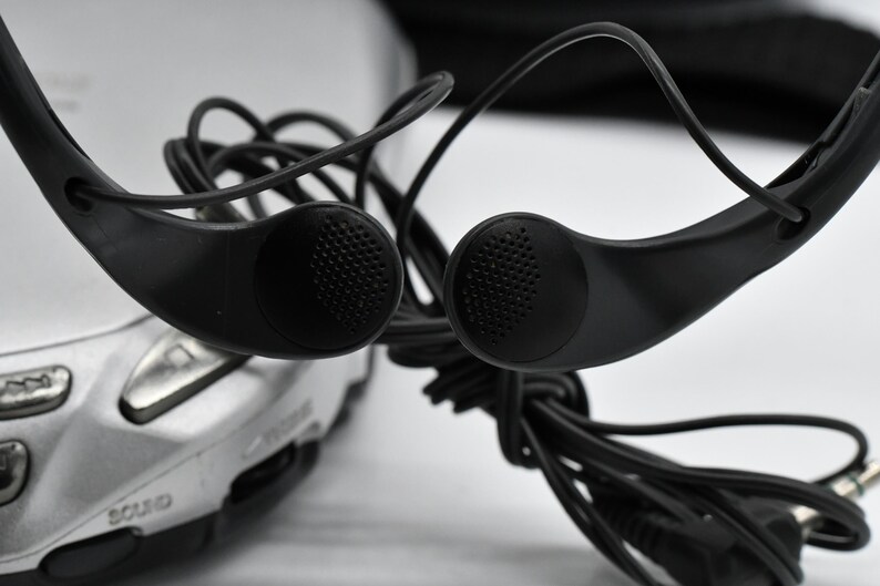 May include: Black over-the-ear headphones with a silver audio device in the background. The headphones have a classic design with ear speakers and a wired connection. The ear speakers have a textured surface.