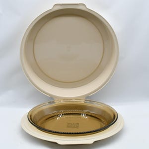 May include: A beige pie carrier with a clear glass pie dish inside. The carrier has a lid that opens to reveal the dish. The pie dish is round and has a slightly tinted color. The carrier is made of plastic.