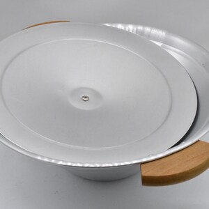 May include: A silver-colored metal bowl with a matching lid, partially open. The bowl has two wooden handles. The lid is slightly ajar, revealing the interior. The bowl is likely used for cooking or serving.