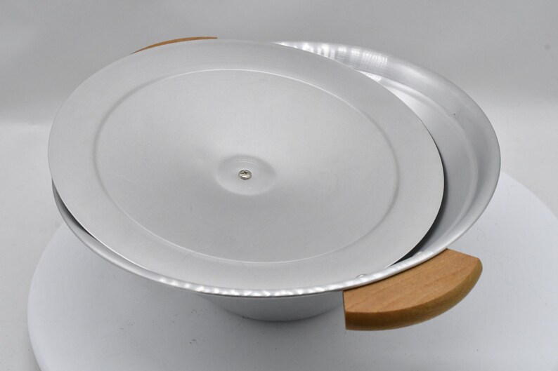 May include: A silver-colored metal bowl with a matching lid, partially open. The bowl has two wooden handles. The lid is slightly ajar, revealing the interior. The bowl is likely used for cooking or serving.